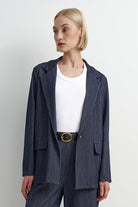 A woman modeling a navy blue pinstripe blazer with matching trousers, styled with a white crewneck top and a gold-buckled belt, against a plain light gray background.