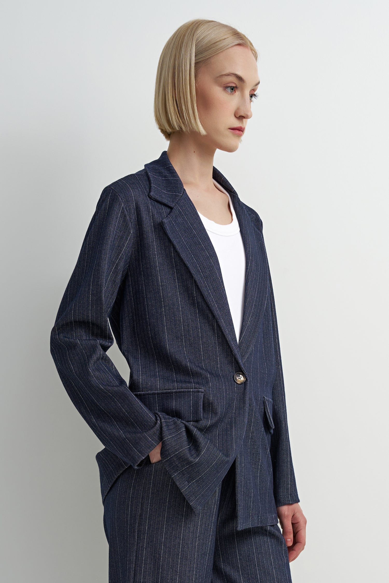 A woman with a short blonde bob is shown in profile wearing a navy blue pinstripe blazer over a white top, paired with matching navy trousers. The photo is shot against a clean, light gray studio background.