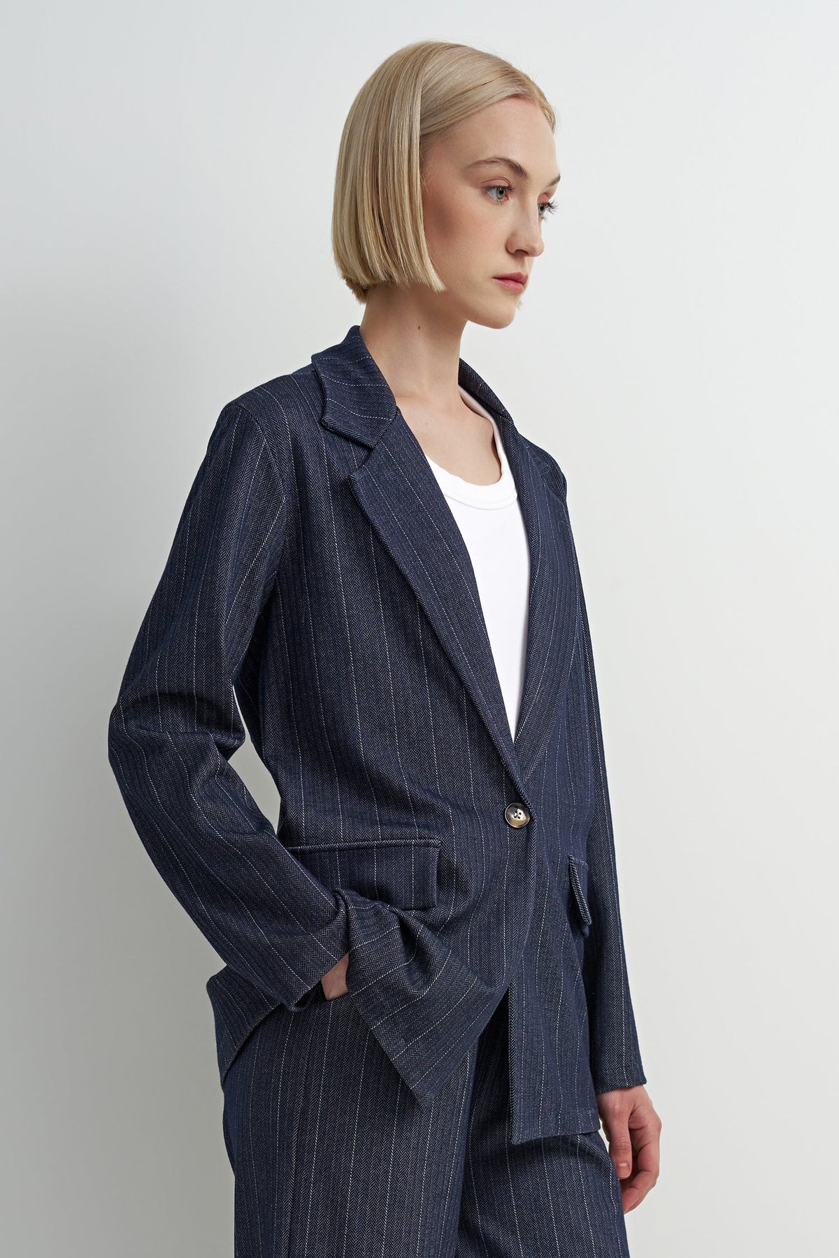 A woman with a short blonde bob is shown in profile wearing a navy blue pinstripe blazer over a white top, paired with matching navy trousers. The photo is shot against a clean, light gray studio background.