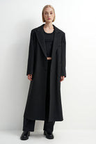 A model stands against a plain white studio background wearing a long black overcoat over a black outfit with black pants and black shoes.