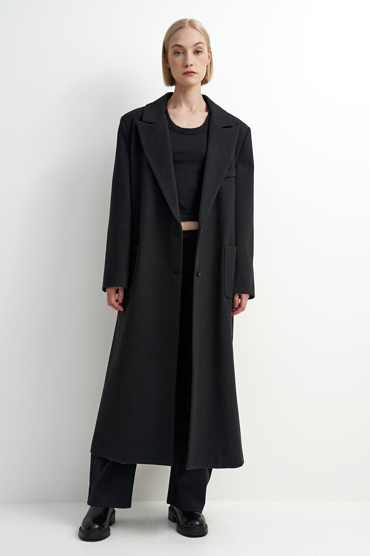 A model stands against a plain white studio background wearing a long black overcoat over a black outfit with black pants and black shoes.