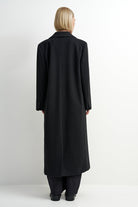 A model viewed from the back wearing a long black coat, black trousers, and black shoes against a plain white studio background.