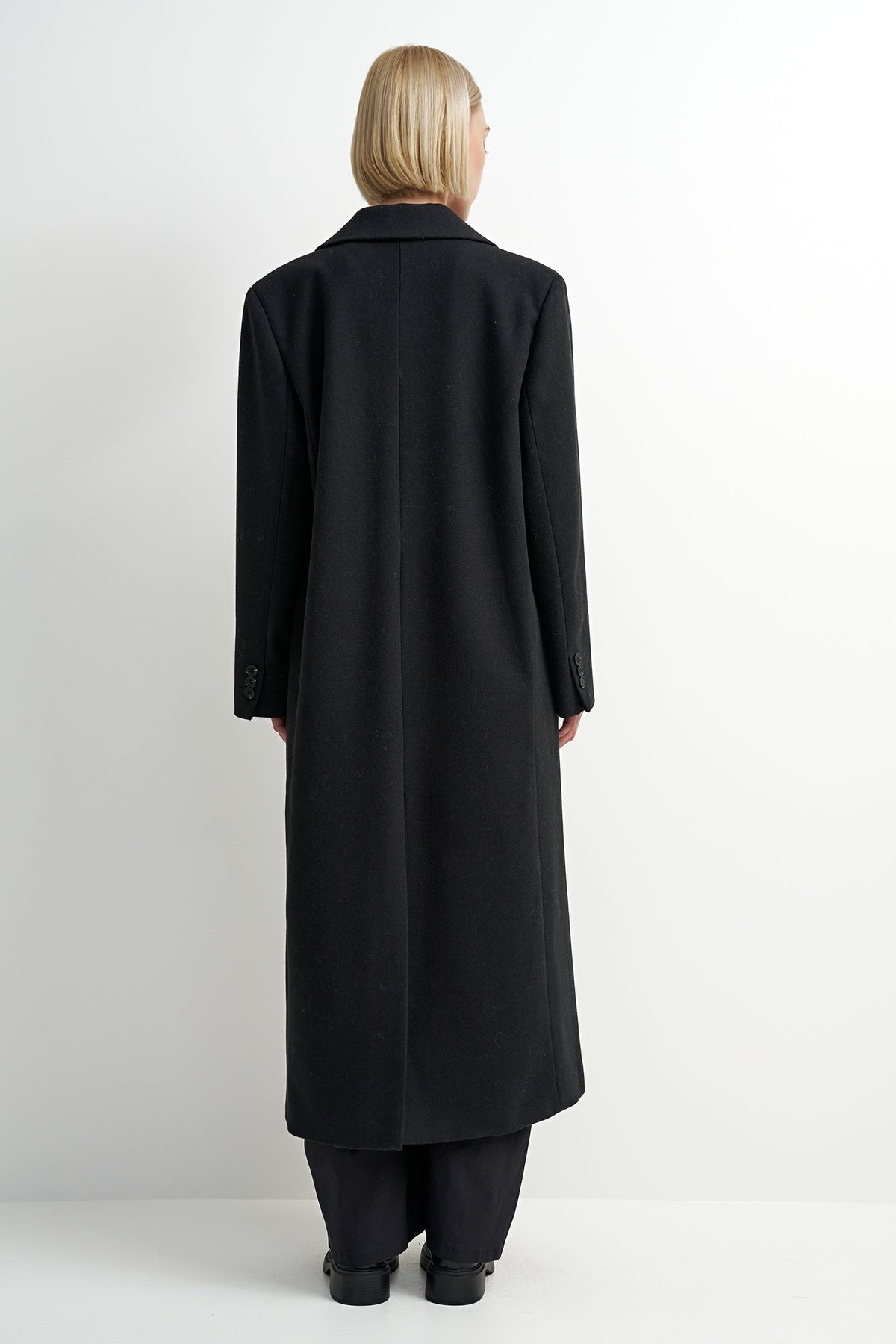 A model viewed from the back wearing a long black coat, black trousers, and black shoes against a plain white studio background.