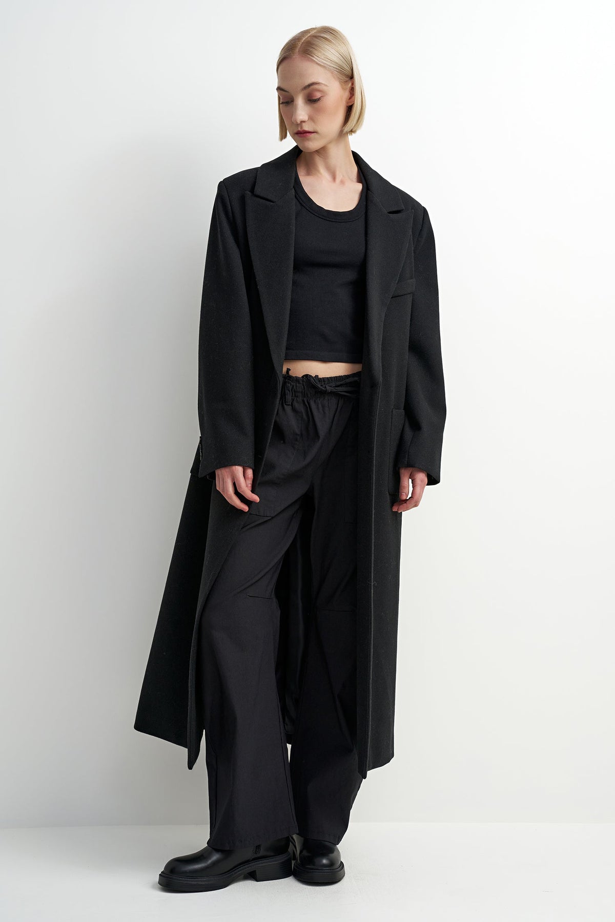A model wearing a long black overcoat over a black crop top, black wide-leg trousers, and black leather shoes, standing against a plain white studio background.