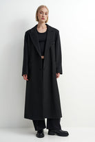 A model wearing a long black wool-blend coat over a black outfit, standing against a plain white studio background.