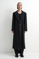 A model stands against a plain white studio background wearing a long black wool-blend coat over a black outfit, with wide-leg trousers and black shoes.