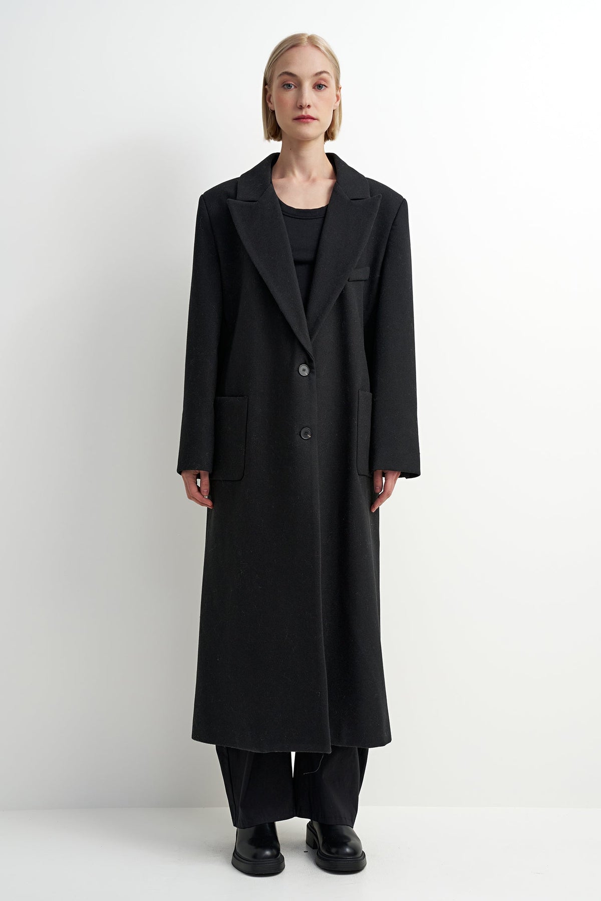 A model stands against a plain white studio background wearing a long black wool-blend coat over a black outfit, with wide-leg trousers and black shoes.