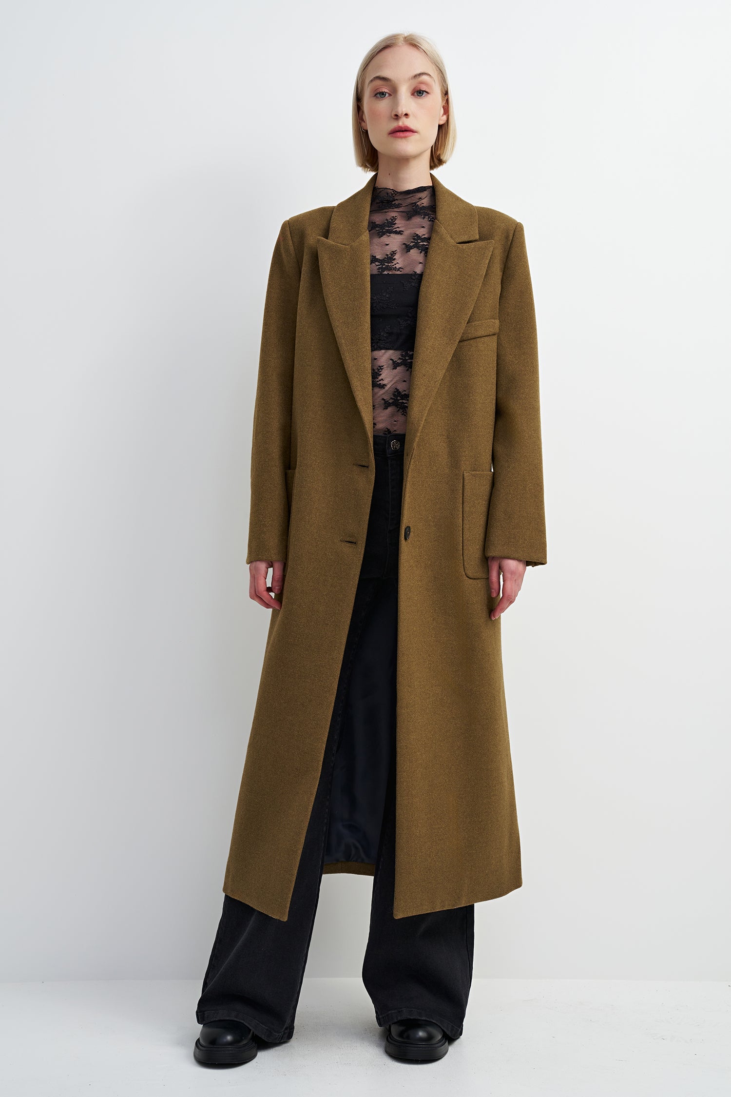 A model stands against a light gray studio background wearing a long, wide lapel camel-brown coat over a black top and black wide-leg trousers.