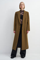 A model stands against a light gray studio background wearing a long, wide lapel camel-brown coat over a black top and black wide-leg trousers.