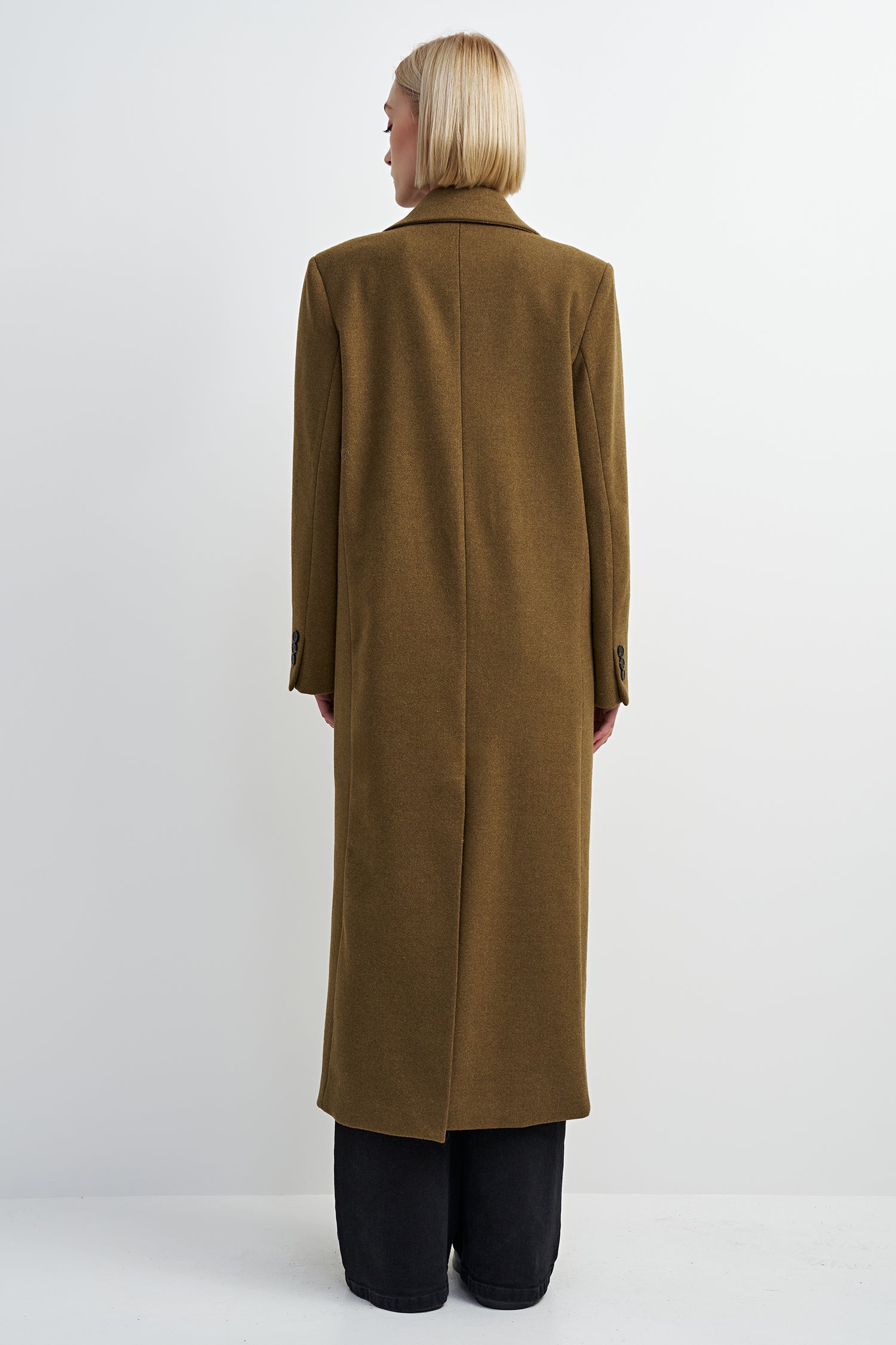 A model viewed from the back wearing a long, straight-line brown coat over black trousers, set against a plain light gray studio background.