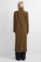A model viewed from the back wearing a long, straight-line brown coat over black trousers, set against a plain light gray studio background.