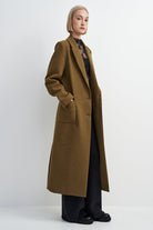 A woman modeling a long, mid-length olive-brown wool coat over a dark patterned top and black wide-leg trousers, standing against a plain light gray studio background.