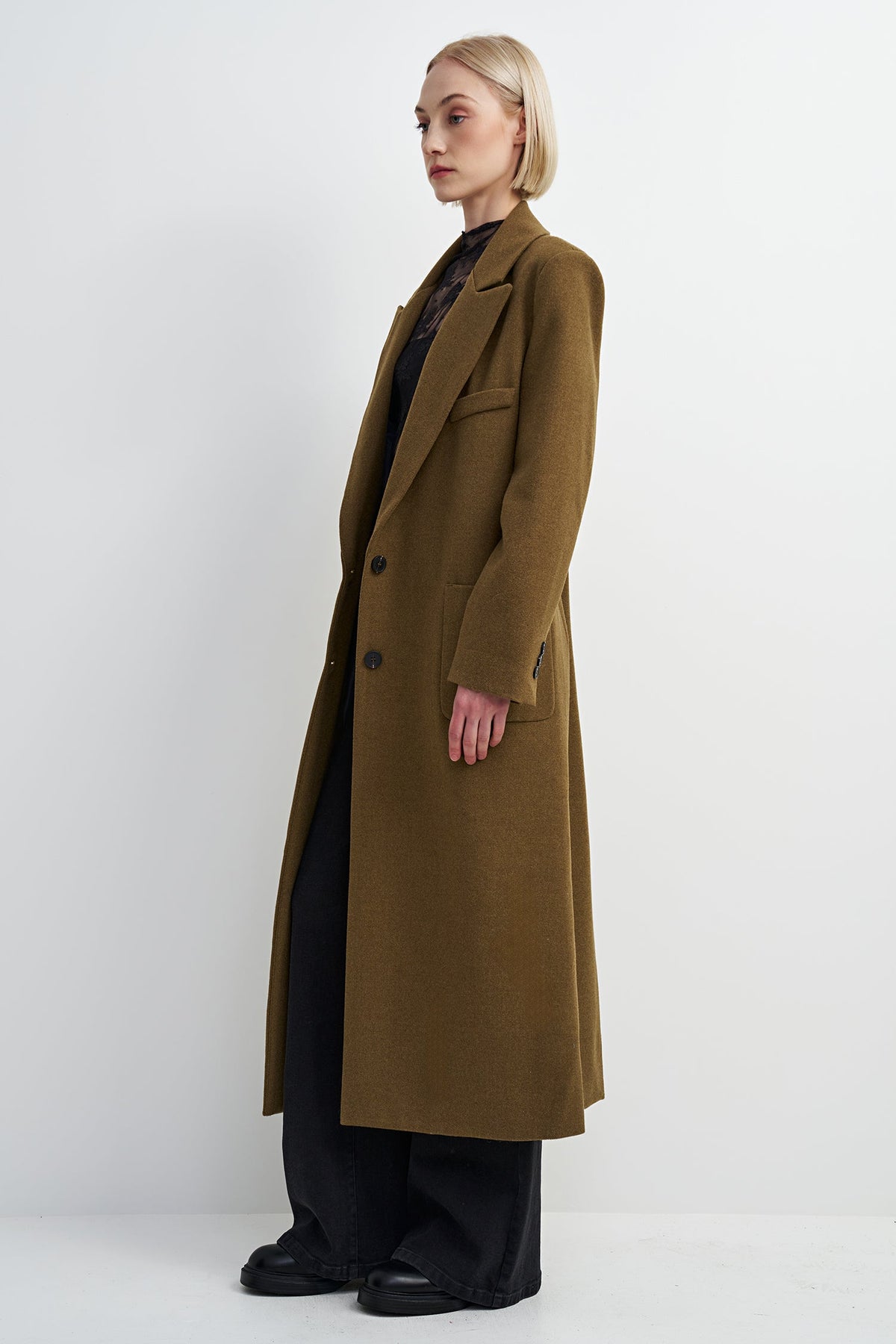 A woman modeling a long, double-breasted camel-brown wool coat over a black top and wide-leg black trousers, photographed against a plain light gray studio background.