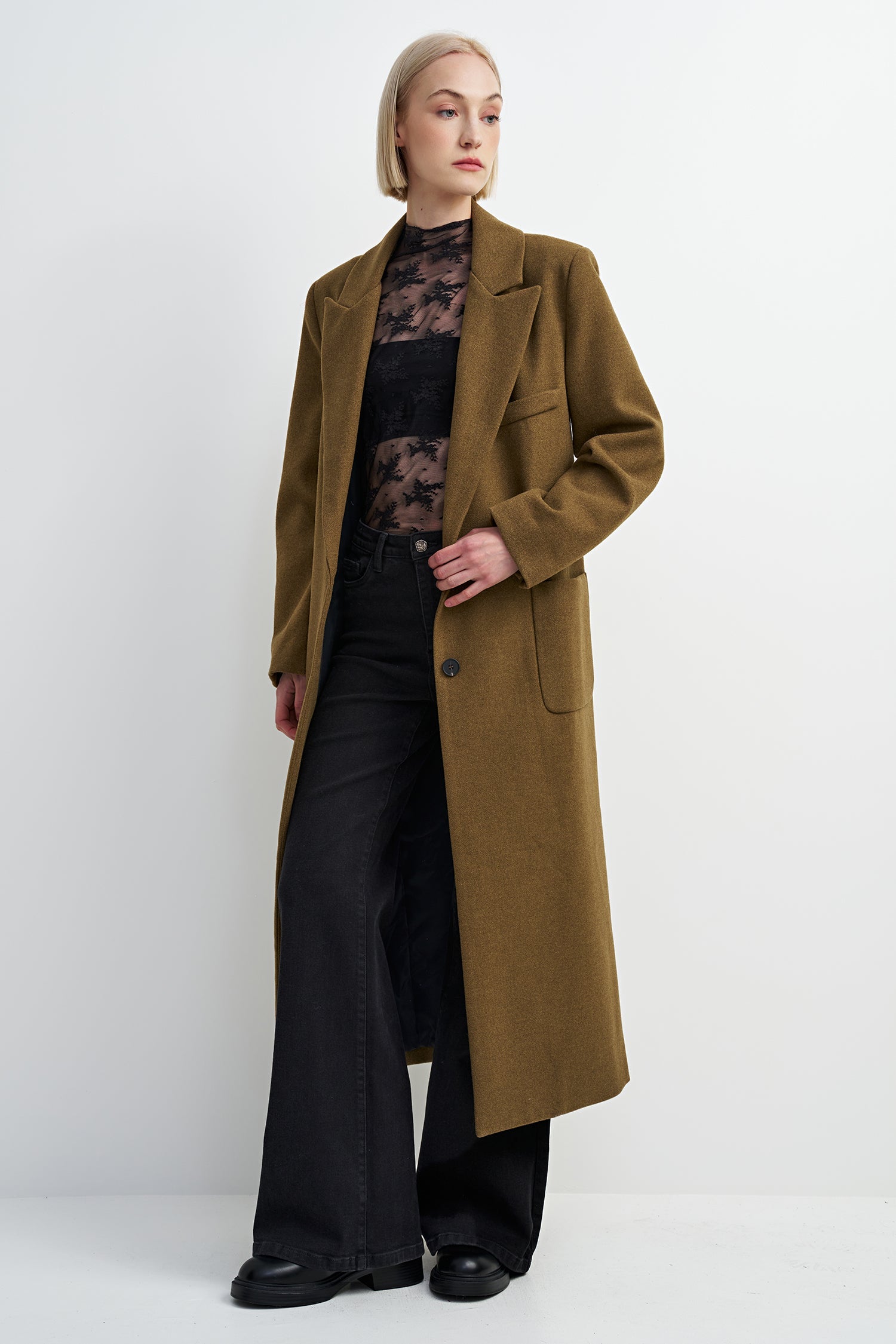 A model wearing a long, olive-brown wool coat over a black lace top and black wide-leg trousers, standing against a plain white studio background.