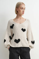 A woman wearing a cream-colored fuzzy knit sweater with black heart motifs and black pants, photographed against a plain light gray studio background.