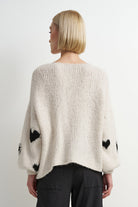 A woman viewed from the back wearing a chunky knit cream sweater with black heart motifs on the sleeves and dark trousers, against a plain light gray studio background.