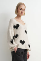 A woman wearing a cream-colored fuzzy knit sweater with black heart motifs, styled with dark gray pants against a plain light gray studio background.