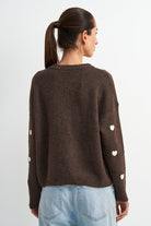 A woman shown from the back wearing a dark brown knit sweater with small white heart details on the sleeves, paired with light blue jeans, against a plain light gray background.