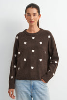 A woman wearing a dark brown knit sweater with small white heart motifs, paired with light blue jeans, standing against a plain light gray background.