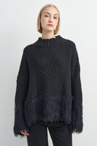 A woman wearing a dark charcoal gray chunky knit sweater with a fuzzy textured hem and flared cuffs, paired with black pants, against a light gray studio background.