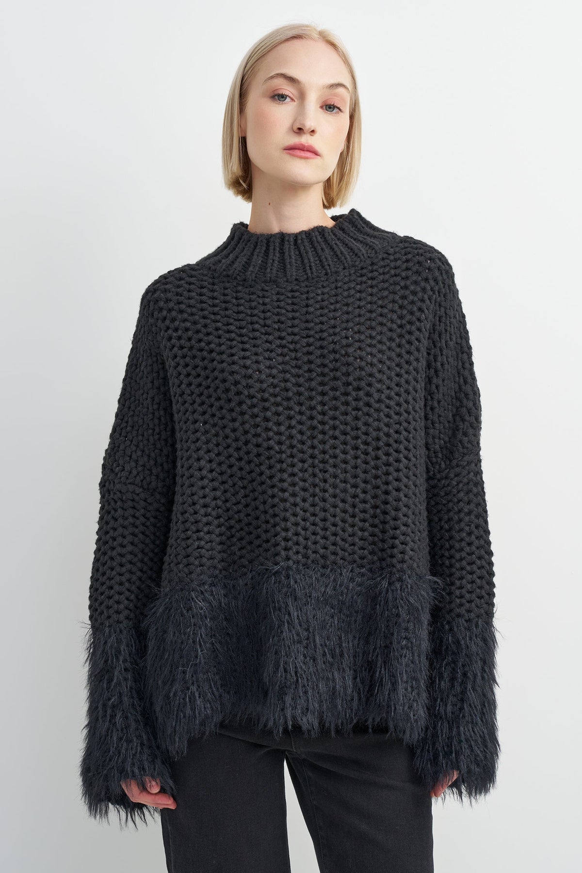 A woman wearing a dark charcoal gray chunky knit sweater with a fuzzy textured hem and flared cuffs, paired with black pants, against a light gray studio background.
