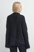 A woman shown from the back wearing a black chunky knit sweater with a fuzzy fringe hem, paired with black pants, against a light gray studio background.