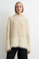 A woman wearing a chunky knit mock neck sweater in cream with a fuzzy fringe hem, paired with black pants, against a plain white studio background.