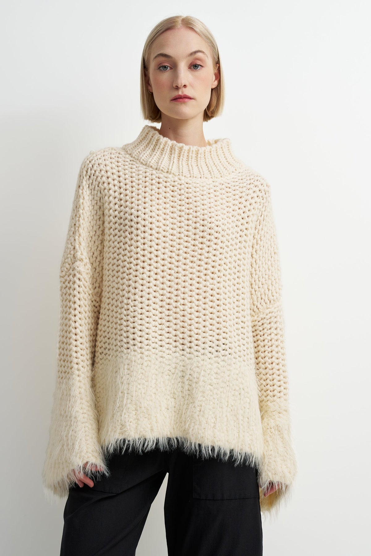 A woman wearing a chunky knit mock neck sweater in cream with a fuzzy fringe hem, paired with black pants, against a plain white studio background.