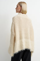 A woman shown from the back wearing a chunky knit open-knit sweater in cream, paired with black pants, against a plain light gray studio background.