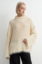 A woman wearing a chunky knit cream-colored sweater with a fuzzy textured hem and wide sleeves, paired with black pants, against a plain light gray background.