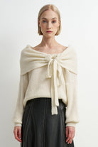 A woman modeling a cream-colored knit sweater with a wide draped collar tied in a large bow at the chest, paired with a black skirt, against a plain light gray studio background.