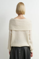 A woman viewed from the back wearing a cream off-the-shoulder ribbed knit sweater and a black skirt, against a plain light gray studio background.