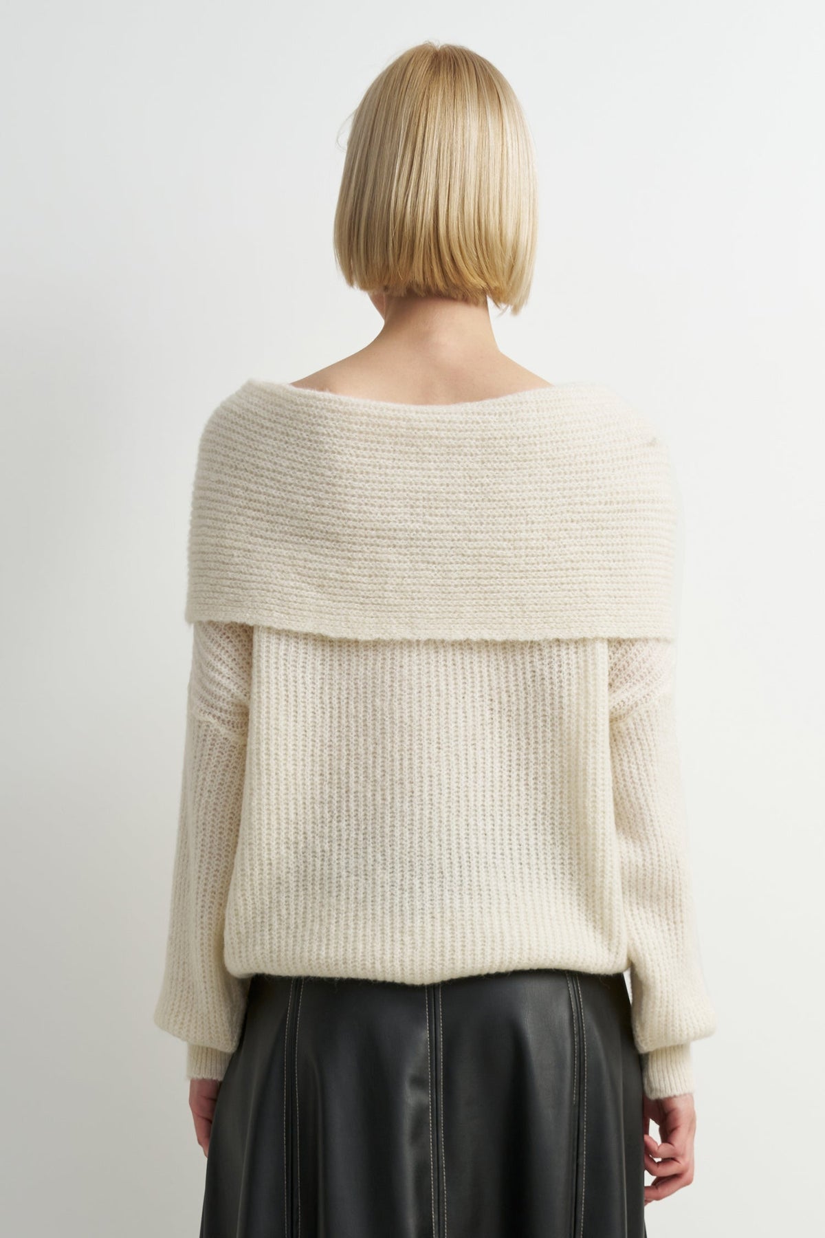 A woman viewed from the back wearing a cream off-the-shoulder ribbed knit sweater and a black skirt, against a plain light gray studio background.