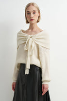 A woman modeling a cream-colored, textured knit sweater with a large front bow detail, paired with a black skirt, against a plain white studio background.