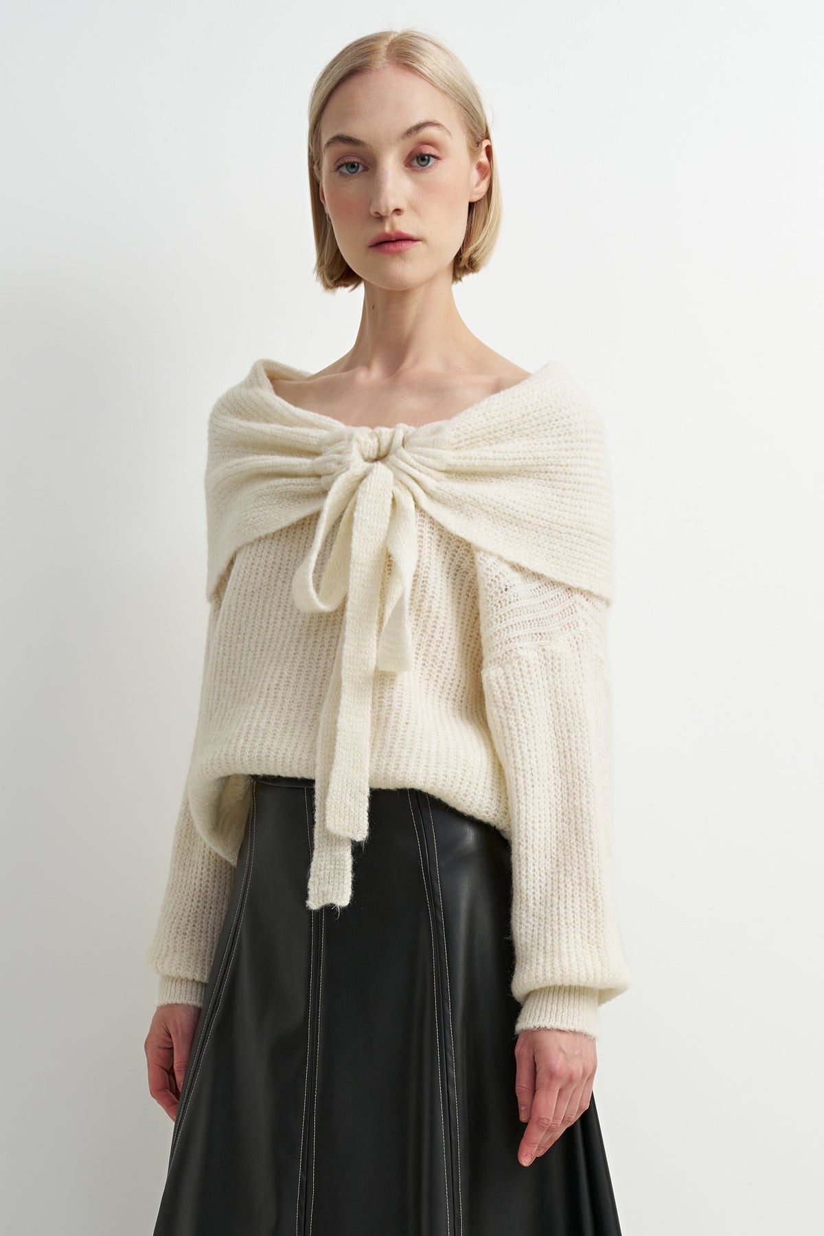 A woman modeling a cream-colored, textured knit sweater with a large front bow detail, paired with a black skirt, against a plain white studio background.