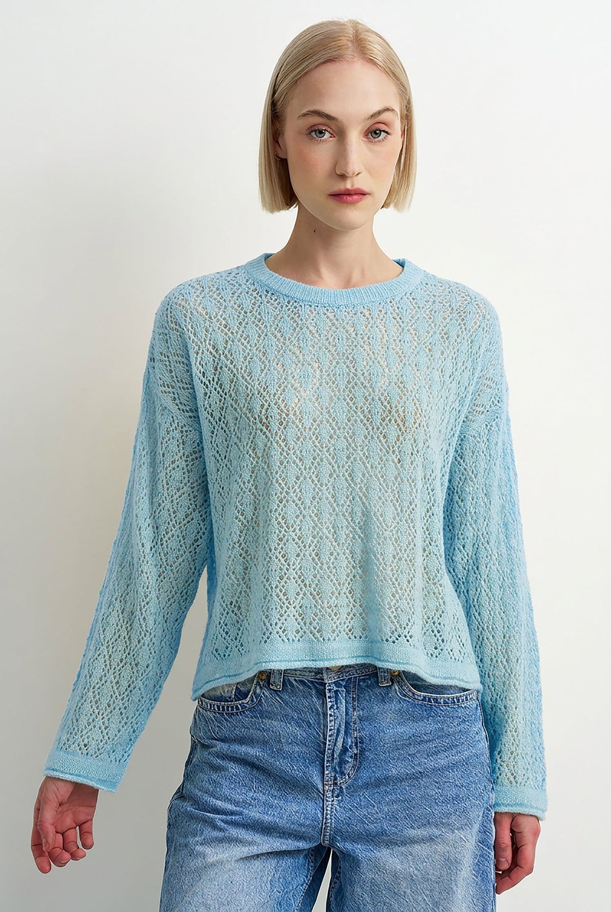 A woman modeling a light blue open-knit long-sleeve sweater with a crew neck, paired with blue jeans, against a plain light gray background.