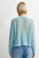 A woman viewed from behind wearing a light blue open-knit cropped sweater and blue jeans against a plain light gray studio background.