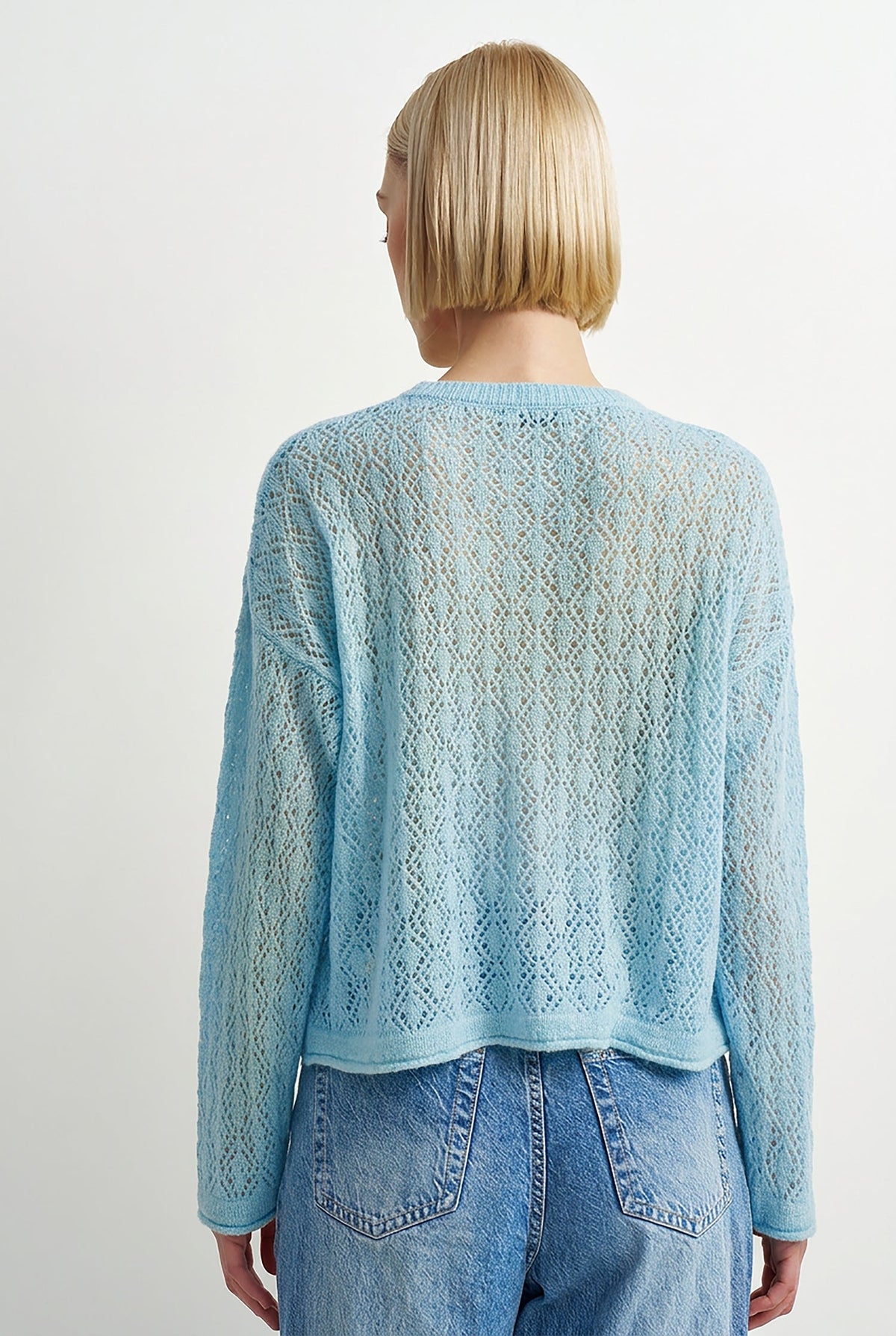 A woman viewed from behind wearing a light blue open-knit cropped sweater and blue jeans against a plain light gray studio background.