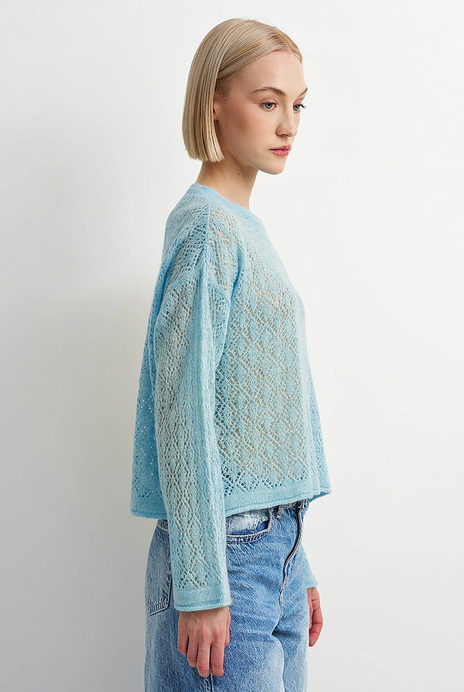 A woman with a short blonde bob is shown in profile wearing a light blue open-knit cropped sweater and light-wash blue jeans against a plain white studio background.