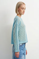 A woman with a short blonde bob is shown in profile wearing a light blue open-knit cropped sweater and light-wash blue jeans against a plain white studio background.