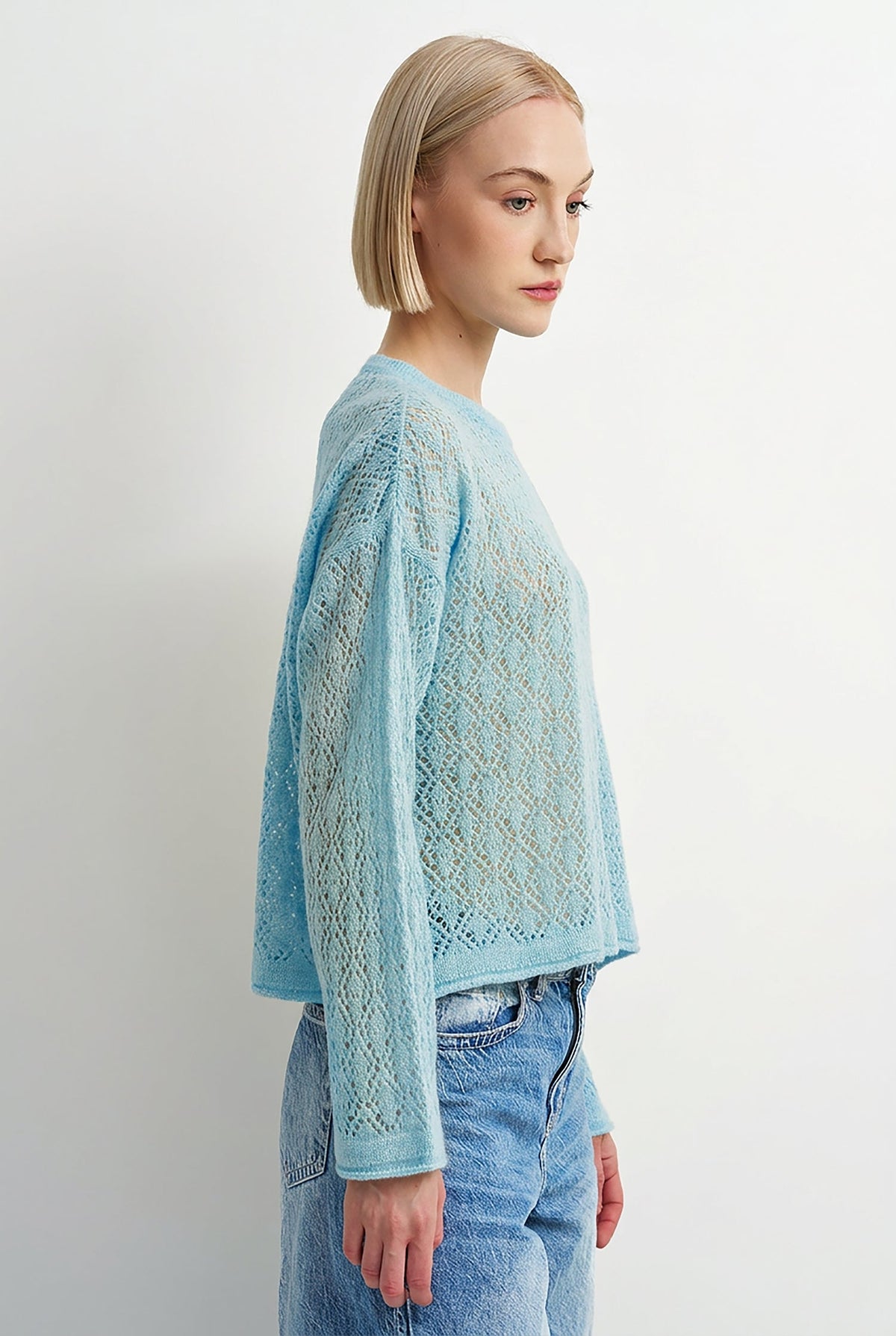 A woman with a short blonde bob is shown in profile wearing a light blue open-knit cropped sweater and light-wash blue jeans against a plain white studio background.