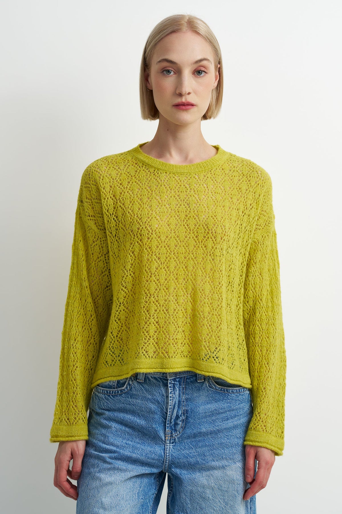 A woman wearing a textured, open-knit long-sleeve sweater in a vibrant yellow-green (lime/chartreuse) color, paired with blue jeans, against a plain light gray studio background.