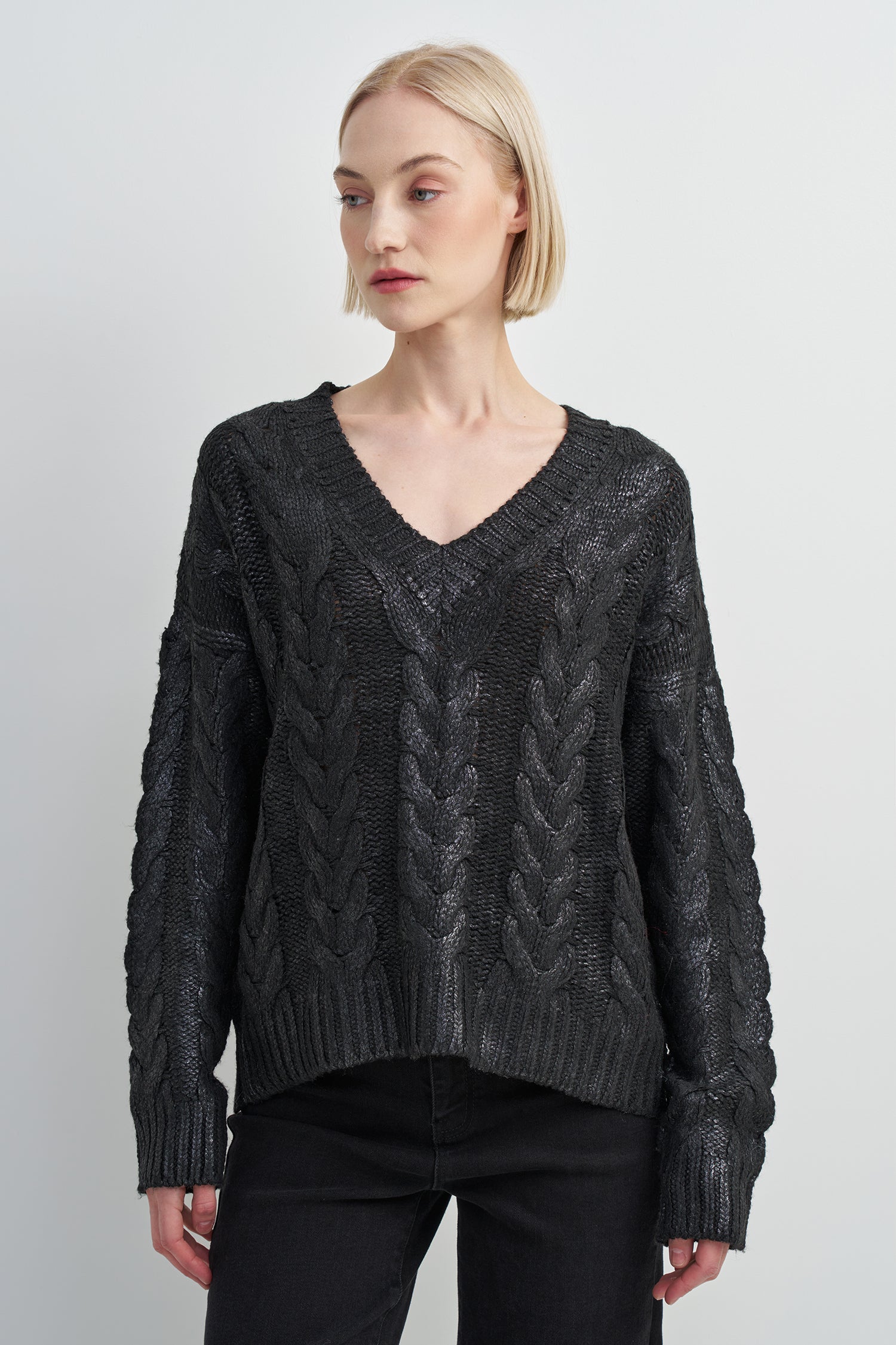 A woman modeling a dark charcoal gray cable-knit sweater with a V-neck and ribbed cuffs, paired with black pants, against a light gray studio background.
