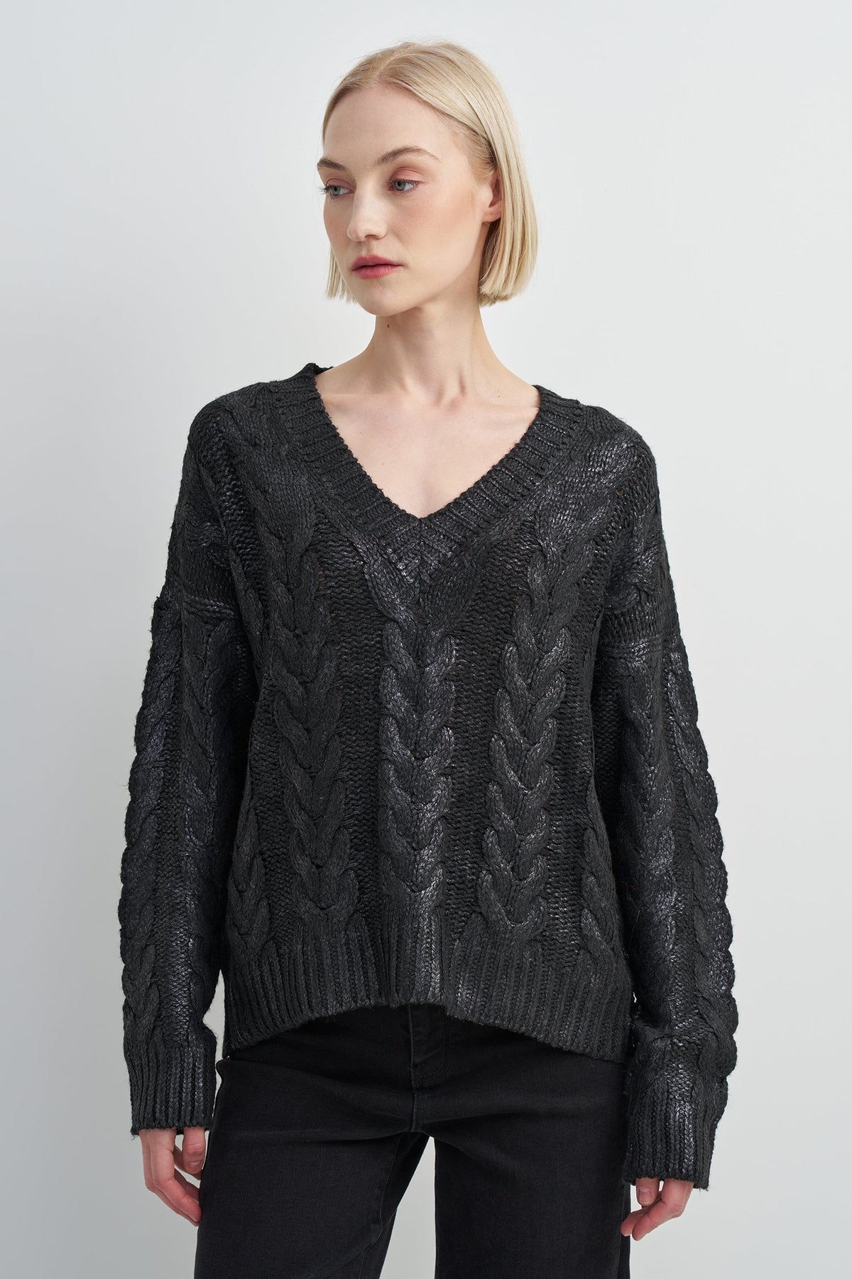 A woman modeling a dark charcoal gray cable-knit sweater with a V-neck and ribbed cuffs, paired with black pants, against a light gray studio background.