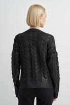 A woman viewed from the back wearing a black cable-knit sweater and black pants against a light gray studio background.