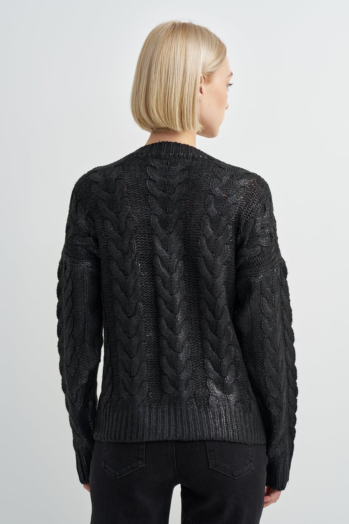 A woman viewed from the back wearing a black cable-knit sweater and black pants against a light gray studio background.