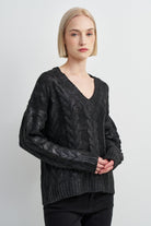 A woman with short blonde hair wearing a dark gray cable-knit V-neck sweater and black pants, standing against a light gray studio background.