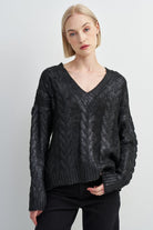 A blonde model wearing a black cable-knit V-neck sweater and black pants, standing against a light gray studio background.