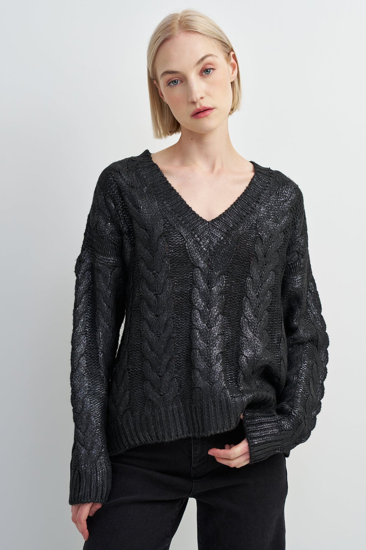 A blonde model wearing a black cable-knit V-neck sweater and black pants, standing against a light gray studio background.