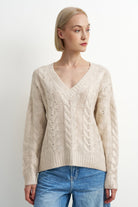 A woman wearing a cream-colored cable-knit V-neck sweater and blue jeans, photographed against a plain light gray studio background.